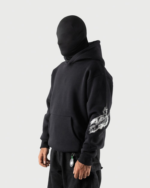 A person wearing a black hooded sweatshirt with a large white graphic print on the left sleeve, paired with black pants, shown against a light gray studio background.