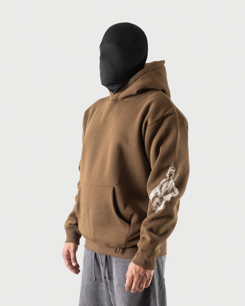 A person wearing a brown pullover hoodie with a white distressed graphic on the left sleeve, styled with gray sweatpants and a black balaclava against a light gray studio background.